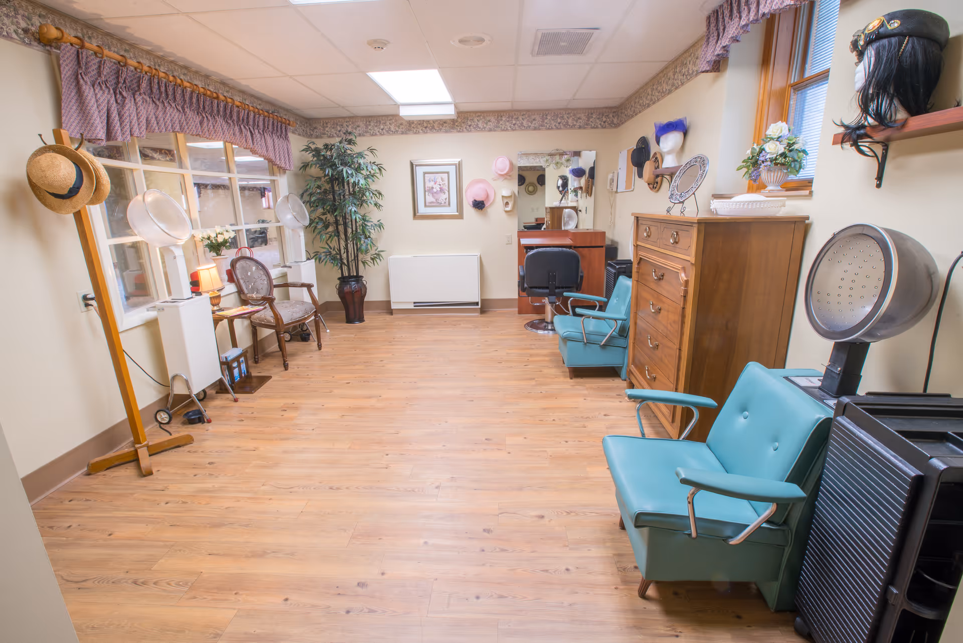 Interior of a vintage-style hair salon room with wooden flooring, teal salon chairs, hair dryers, a wooden dresser, and hats hanging on the walls. There is a large mirror on the far wall with various hats displayed above it, a potted plant in the corner, and a window with a purple valance curtain.