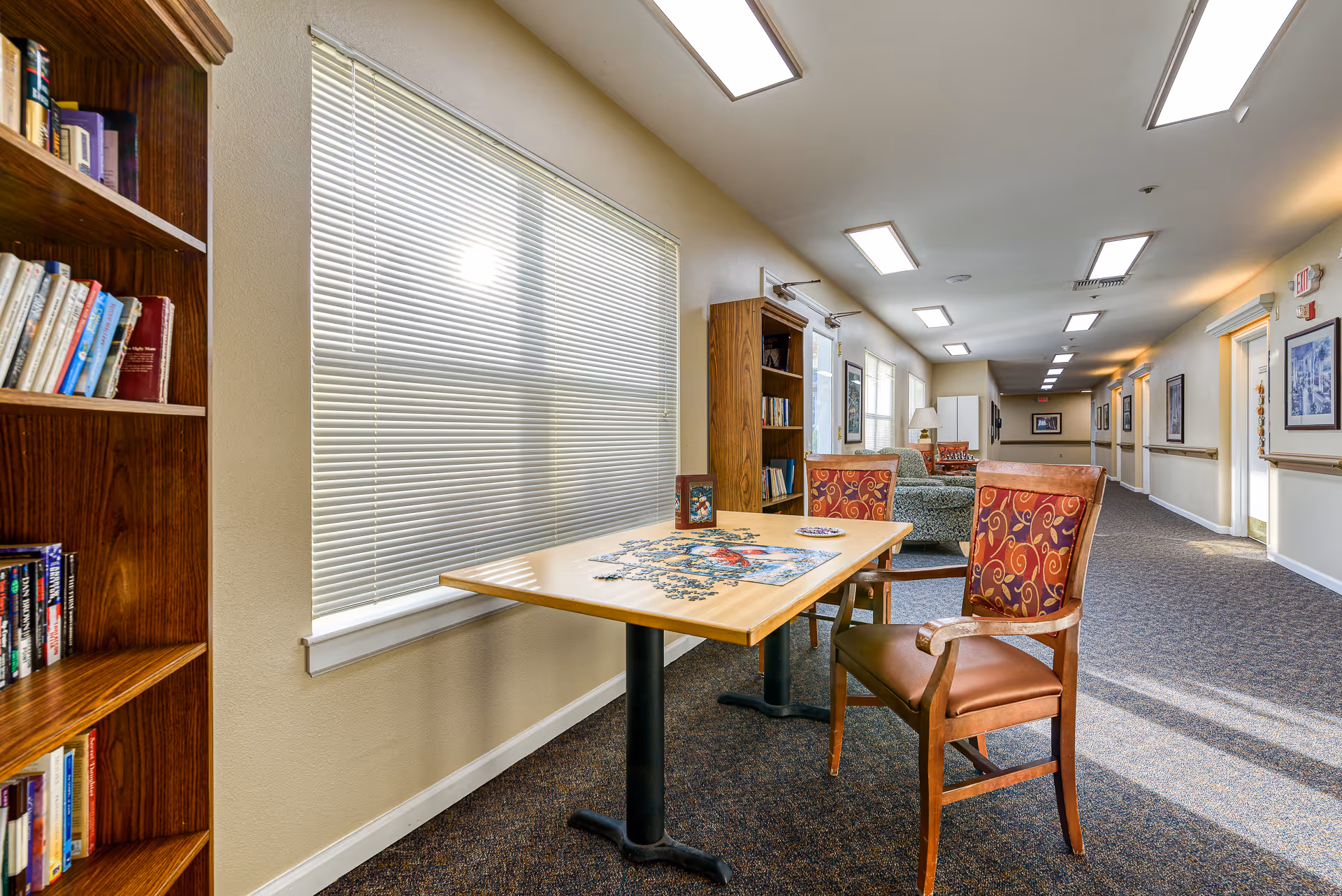 A well-lit hallway in a senior living facility with a table and two chairs in the foreground. The table has a partially completed jigsaw puzzle on it. There are bookshelves filled with books along the wall, windows with blinds, and comfortable seating further down the hallway. The walls are decorated with framed pictures and the floor is carpeted.