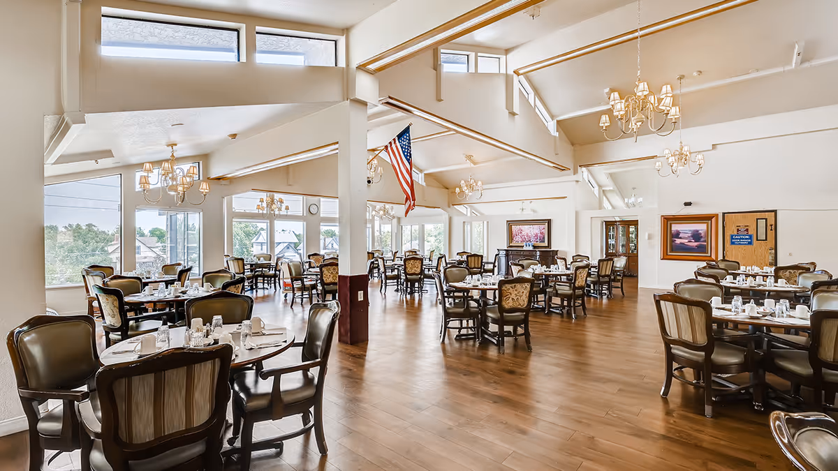 Spacious dining room with multiple round tables set with cups, glasses, and napkins. The room features large windows allowing natural light, chandeliers hanging from a high ceiling, wooden flooring, and an American flag hanging from a beam. There are framed paintings on the walls and a door with a caution sign.