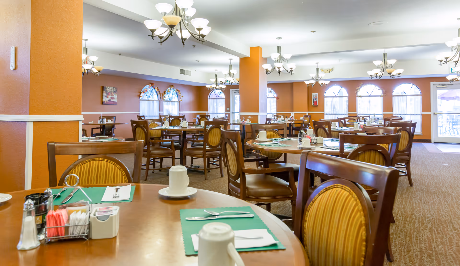 A spacious dining room with multiple round wooden tables and chairs arranged neatly. Each table is set with placemats, cups, utensils, and condiments. The room has warm orange walls, large arched windows with curtains, and several ceiling light fixtures providing bright illumination.
