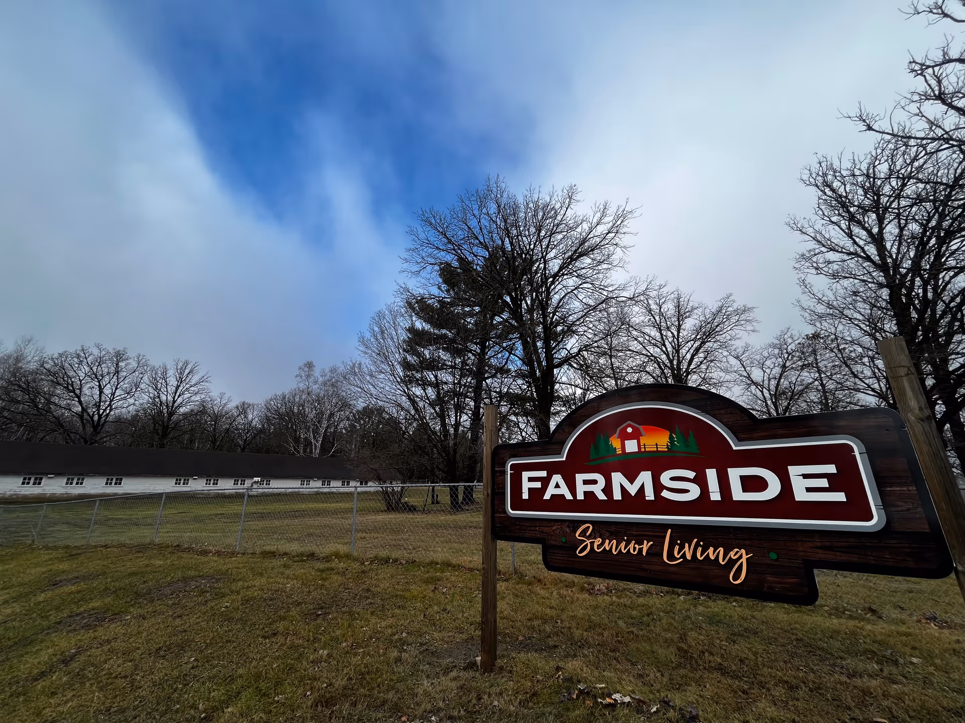 A wooden sign that reads 'Farmside Senior Living' stands in front of a grassy yard with trees and a long white building under a cloudy sky.