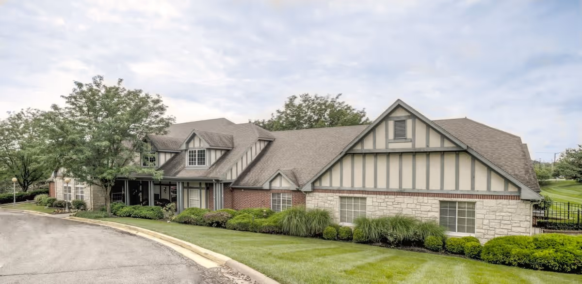 Exterior view of a single-story building with a combination of brick, stone, and wood paneling. The building has a sloped roof with dormer windows and is surrounded by well-maintained green grass, shrubs, and trees. A paved driveway curves along the front of the building under a partly cloudy sky.
