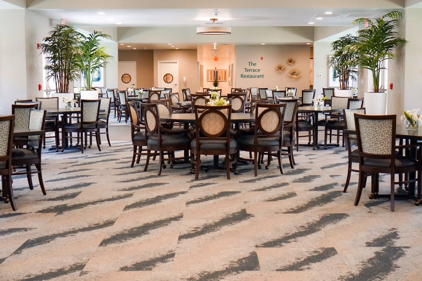 A spacious dining room with multiple round and square tables surrounded by chairs. The room has a patterned carpet and large potted plants on either side. On the back wall, there is a sign that reads 'The Terrace Restaurant'. The room is well-lit with ceiling lights.