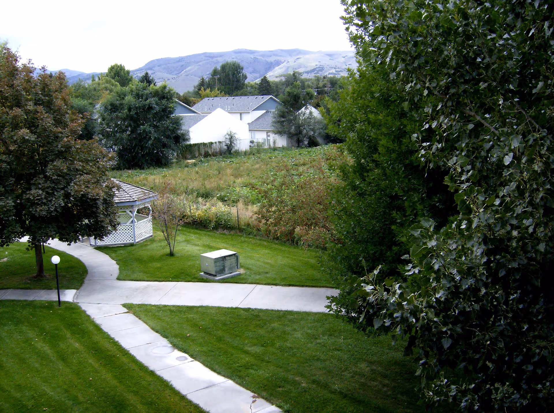 A landscaped outdoor area with green grass, a paved walkway, a white gazebo, and various trees and bushes. In the background, there are residential houses and hills under a cloudy sky.