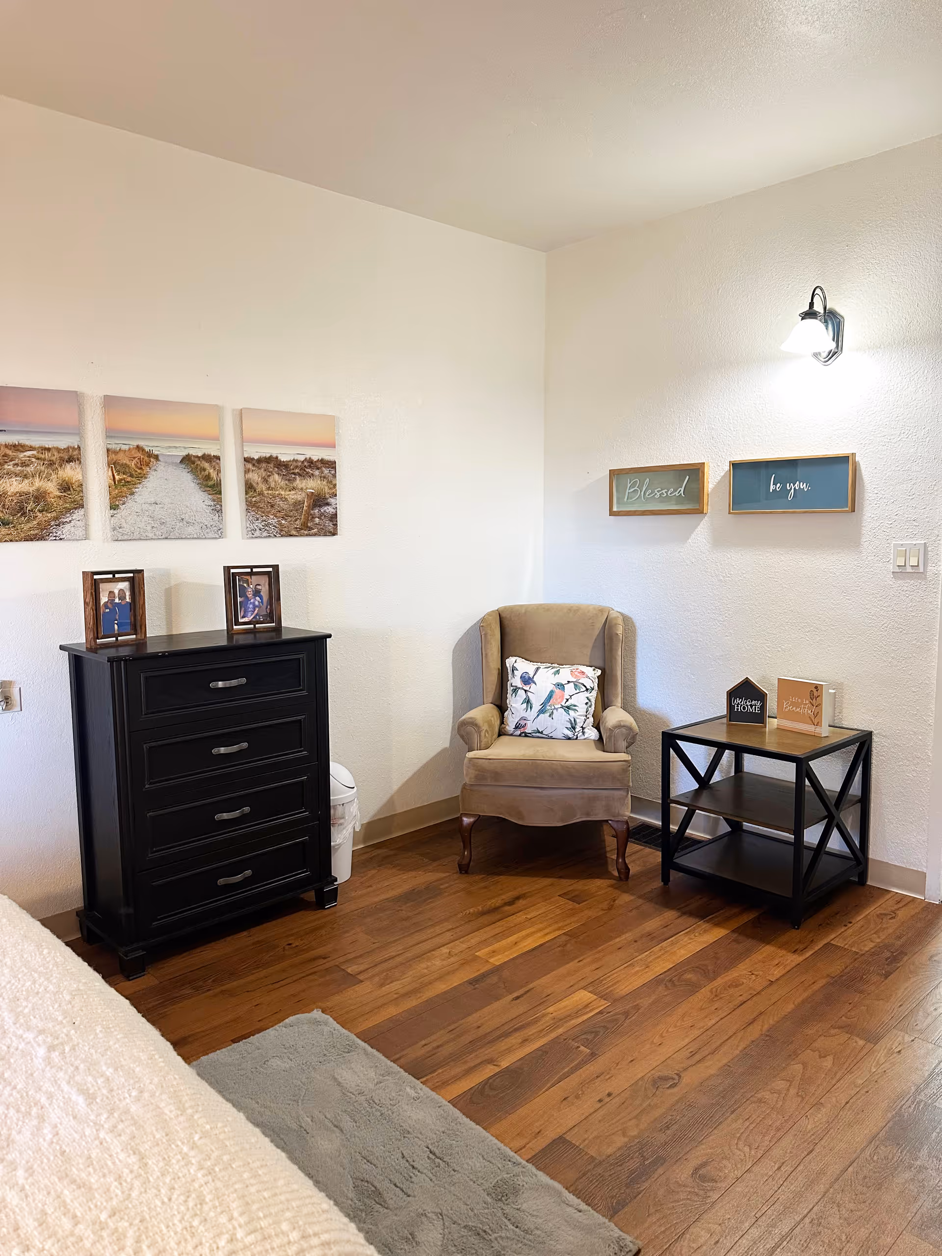 Cozy bedroom corner with an upholstered armchair, black dresser, nightstand, wall art, and hardwood floors.