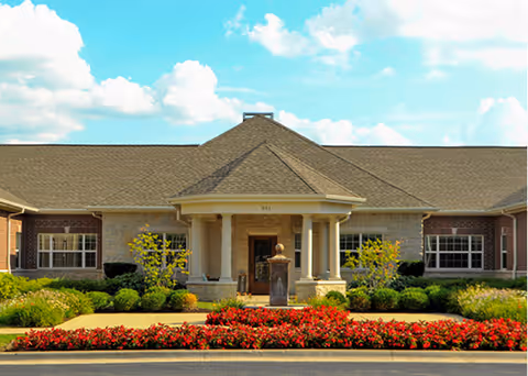 Front exterior view of a single-story brick building with a peaked roof and a covered entrance supported by columns. The building is surrounded by well-maintained landscaping including bushes, small trees, and a flower bed with red flowers in front.