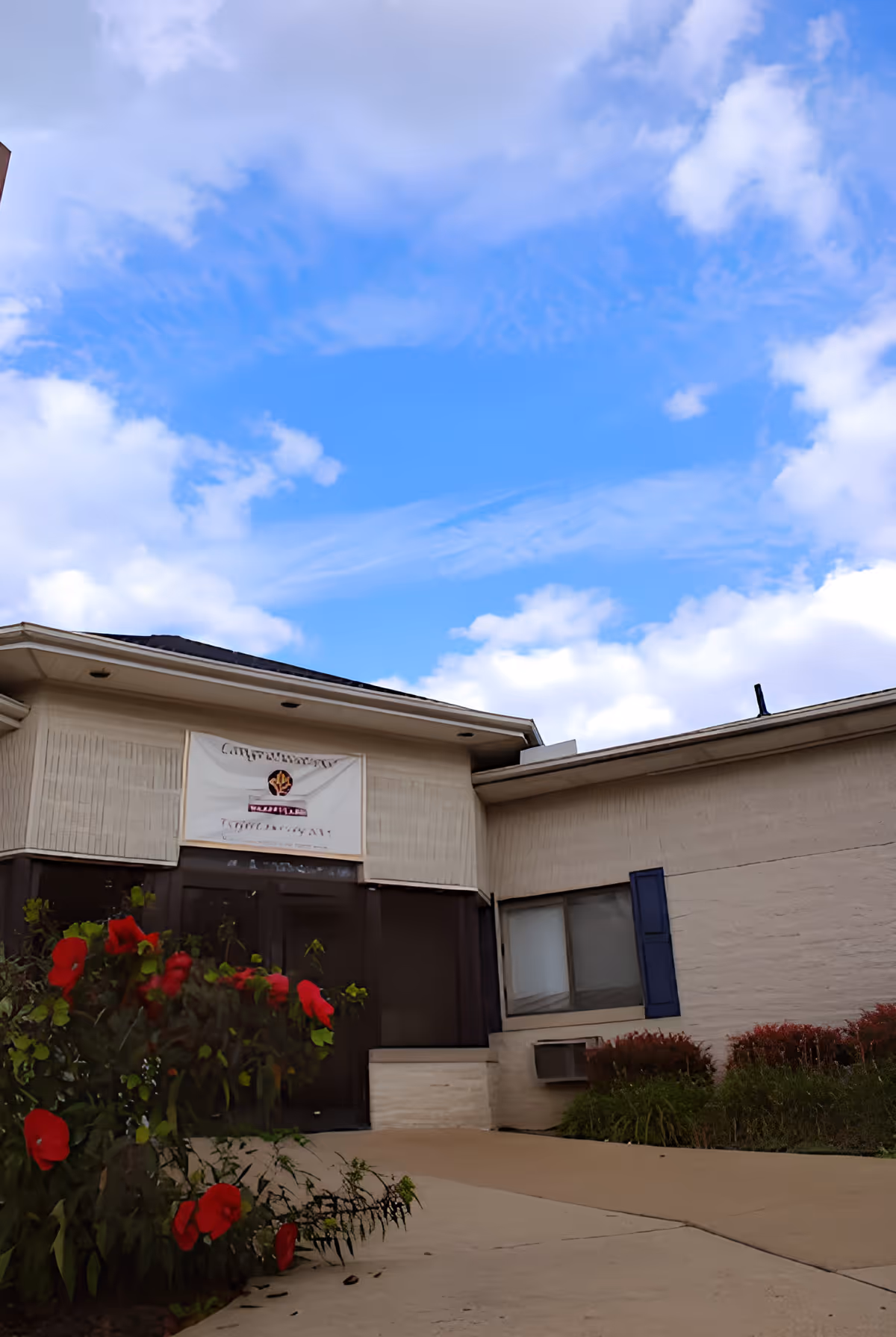 Exterior view of Crestmont North Nursing facility showing the entrance with a beige building, a window with blue shutters, and red flowers in the foreground under a partly cloudy blue sky.