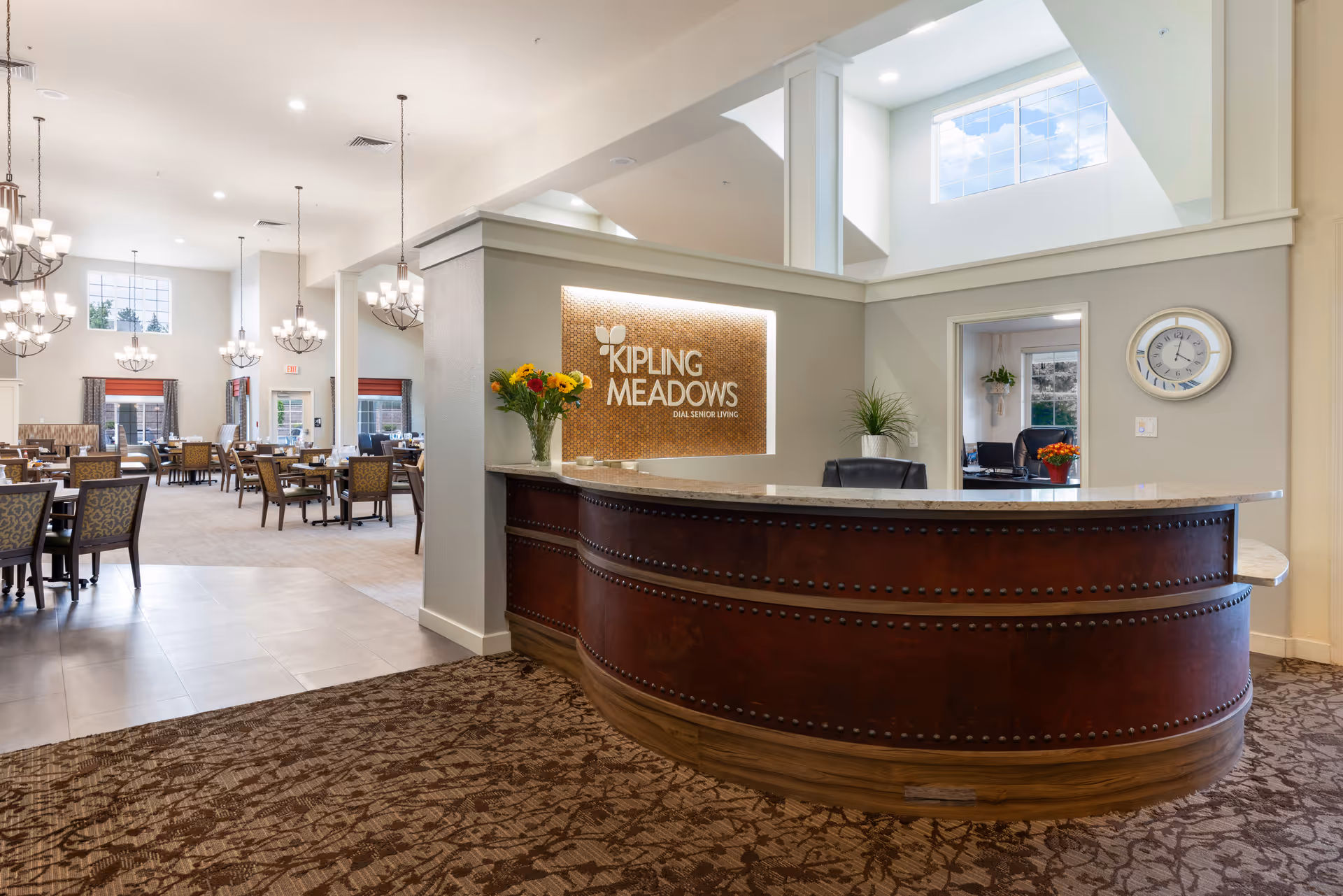 Reception area of Kipling Meadows Senior Living with a curved wooden front desk, a vase of flowers, and a sign on the wall behind the desk. To the left is a spacious dining area with multiple tables and chairs, large windows, and hanging chandeliers providing bright lighting.
