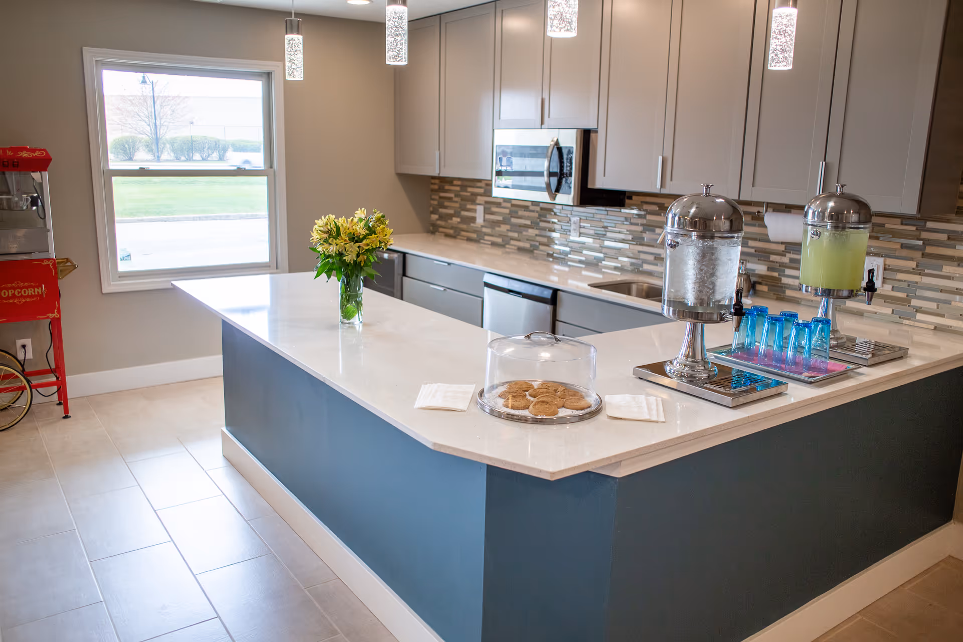 Modern communal kitchen with a large island featuring a vase of flowers, a covered plate of cookies, beverage dispensers and cabinetry in the background.