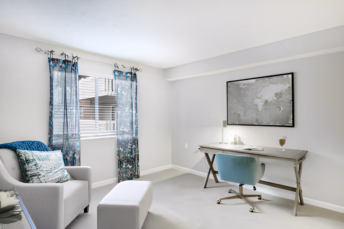 Bright living-room style home office with a desk and blue swivel chair, an armchair and ottoman, and a window with patterned blue curtains.