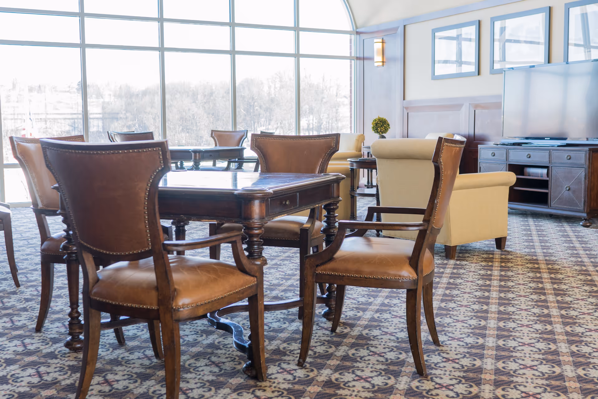 A bright senior living common area with large windows letting in natural light. The room features a wooden table surrounded by leather chairs, beige upholstered armchairs, a patterned carpet, a wooden TV stand with a flat-screen TV, and framed artwork on the wall.