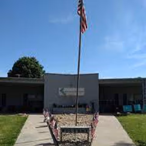 Front entrance of a single-story care facility with a tall American flag pole and small flags lining the walkway.