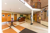 Bright nursing facility lobby with a reception desk, side table with flowers, staircase to an upper level, and double doors.