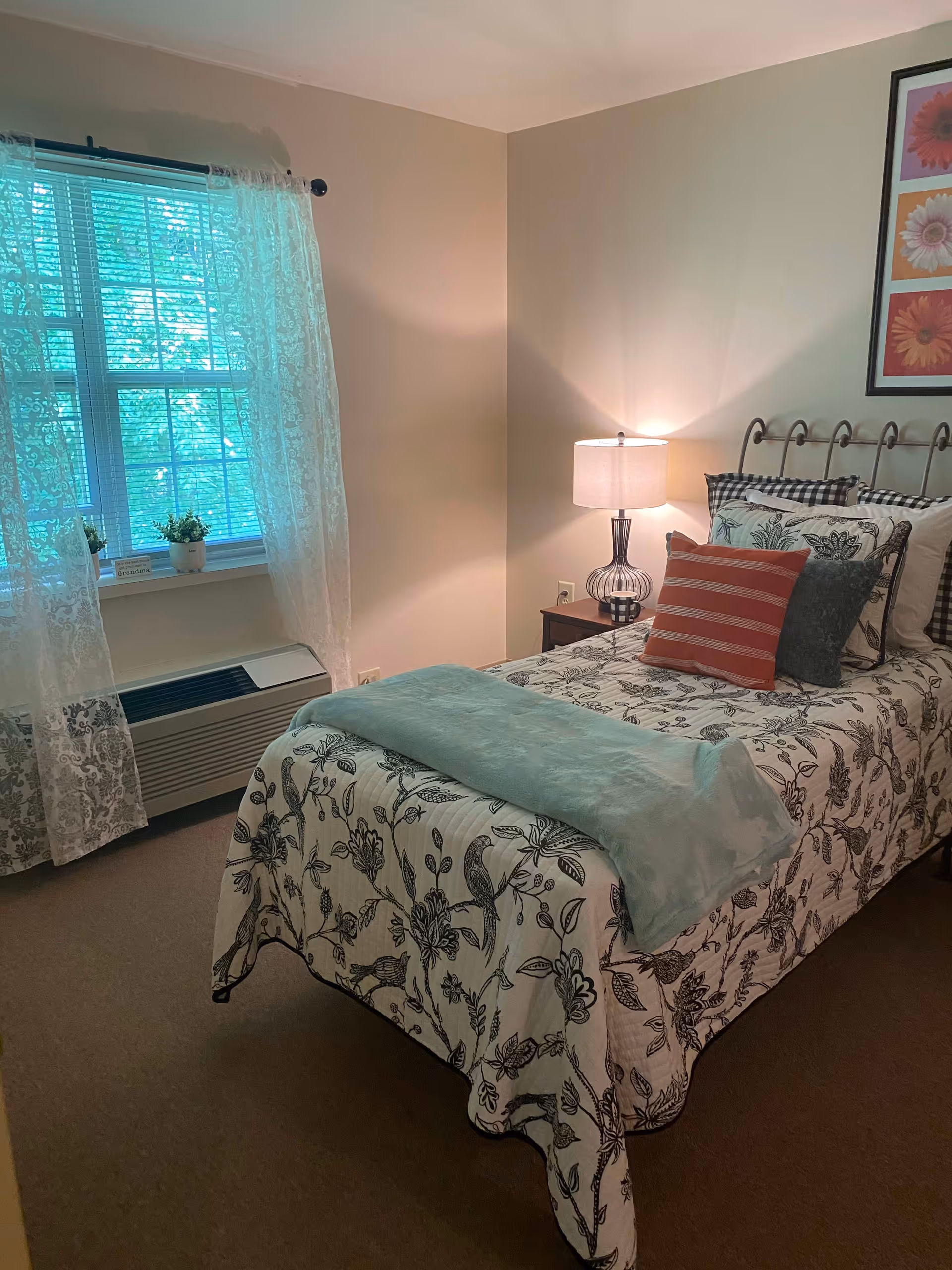 A cozy bedroom with a single bed covered in a floral patterned bedspread and multiple pillows, including a red striped one. A bedside table with a lamp is next to the bed. There is a window with sheer lace curtains letting in natural light, and a framed floral artwork hangs on the wall above the bed.