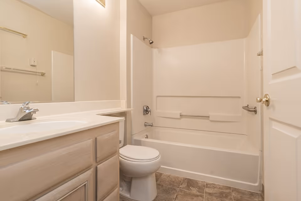 A clean bathroom with a white bathtub and shower combination, a white toilet, a light wood vanity with a white countertop and sink, a large mirror above the sink, and beige walls and floor tiles.