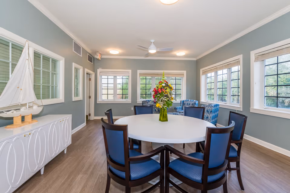 A bright room with a round white table surrounded by six blue cushioned chairs. A vase with colorful flowers is placed in the center of the table. The room has multiple windows with white frames letting in natural light, light blue walls, and a wooden floor. There is a white cabinet with a decorative sailboat on top along one wall and two blue patterned armchairs near the windows.