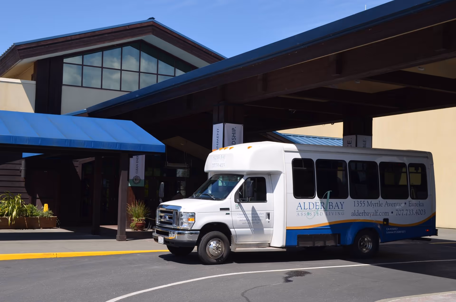 A white and blue shuttle bus parked in front of the entrance to Alder Bay Assisted Living facility. The building has a covered drop-off area with a blue awning and large windows above the entrance.