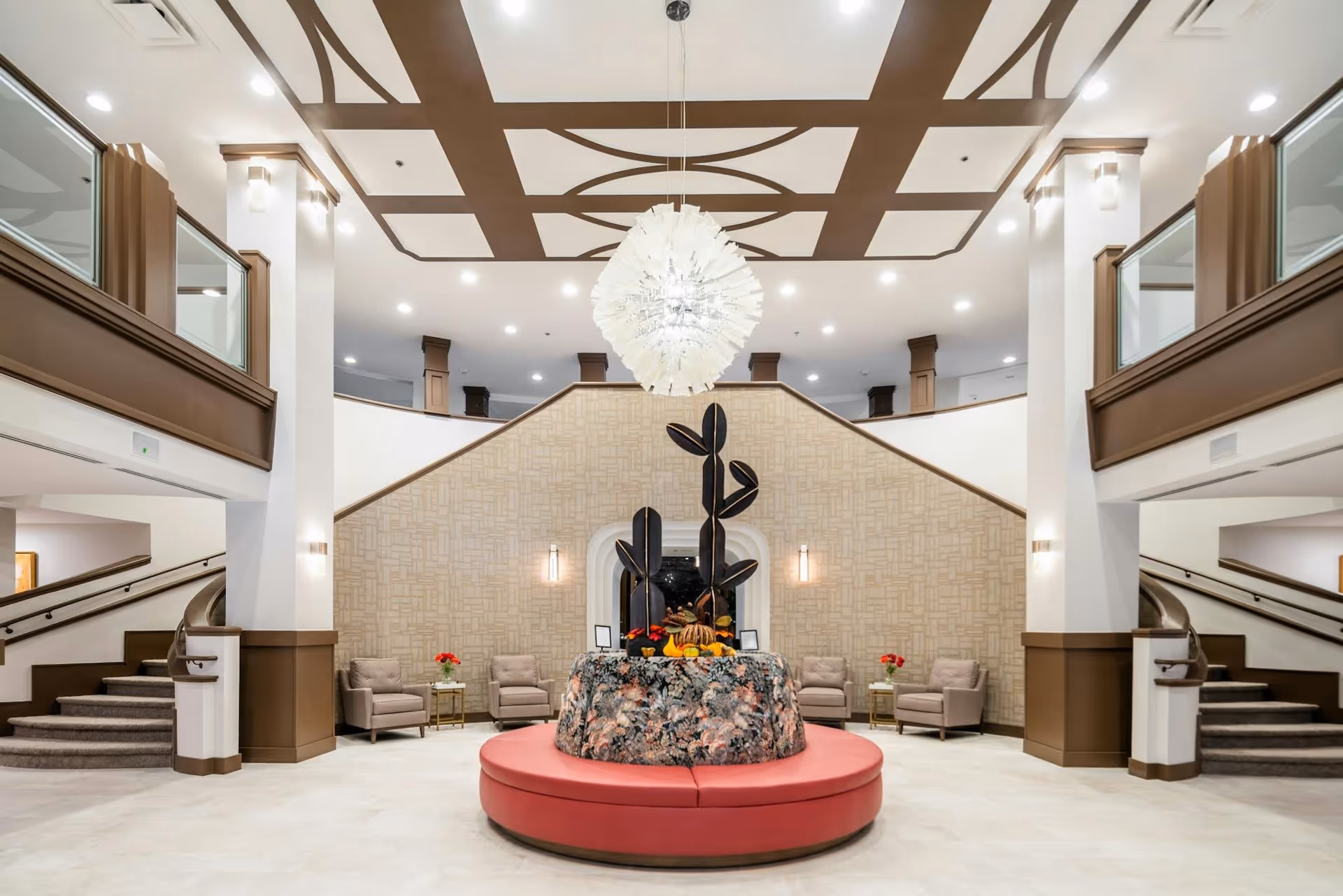 A spacious and elegant lobby area with a central circular seating arrangement upholstered in red, topped with a decorative floral arrangement and black leaf sculptures. The room features a high ceiling with a modern chandelier, patterned ceiling design, and a beige textured wall with two wall sconces. There are two symmetrical staircases on either side leading to an upper level with glass railings and brown trim. Several beige armchairs with small side tables and red flower vases are arranged along the back wall.