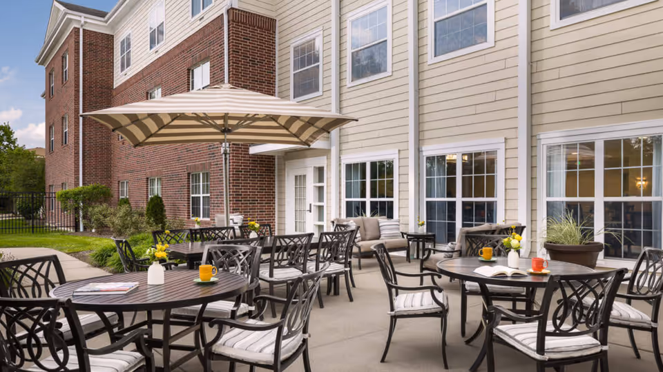 Outdoor patio area at Belmont Village Senior Living Geneva Road featuring multiple round tables with black metal chairs that have white cushions. Some tables have colorful mugs, books, and small flower vases. A large beige and white striped umbrella provides shade over one table. The patio is adjacent to a building with beige siding and large windows.