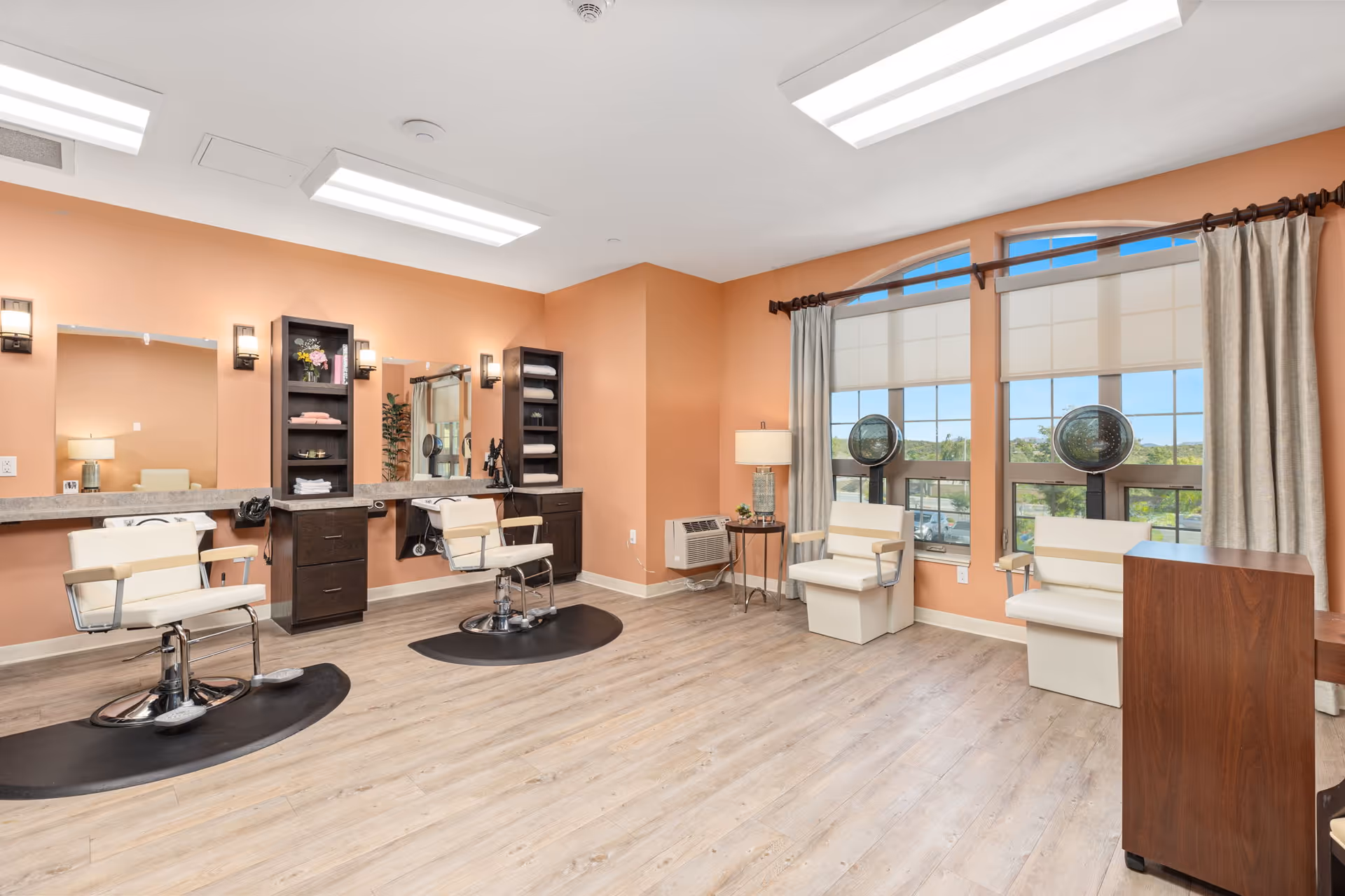 Interior of a hair salon with peach-colored walls, two styling chairs in front of mirrors with shelves holding towels and hair products, two white chairs under hair dryers near large windows with beige curtains, and a small table with a lamp.