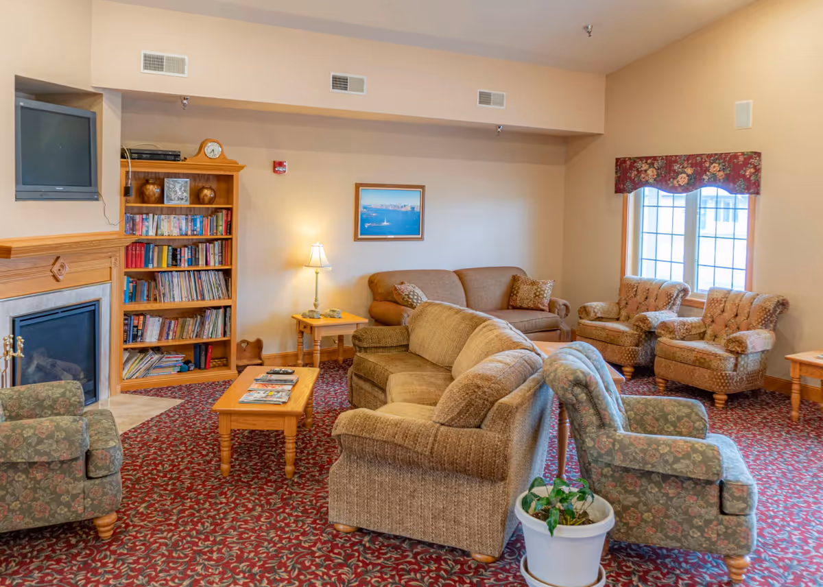 A cozy senior living facility common area with multiple upholstered armchairs and sofas arranged around a wooden coffee table. There is a wooden bookshelf filled with books and decorative items, a fireplace with a mounted TV above it, a side table with a lamp, a framed picture on the wall, and a window with floral valance curtains. The room has a patterned red carpet and beige walls.