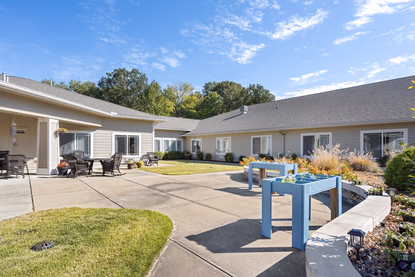 Outdoor courtyard area of Quail Ridge Assisted Living & Memory Care featuring a paved walkway, green grass patches, two blue raised garden beds, patio tables and chairs, surrounded by a single-story building with windows and a clear blue sky above.