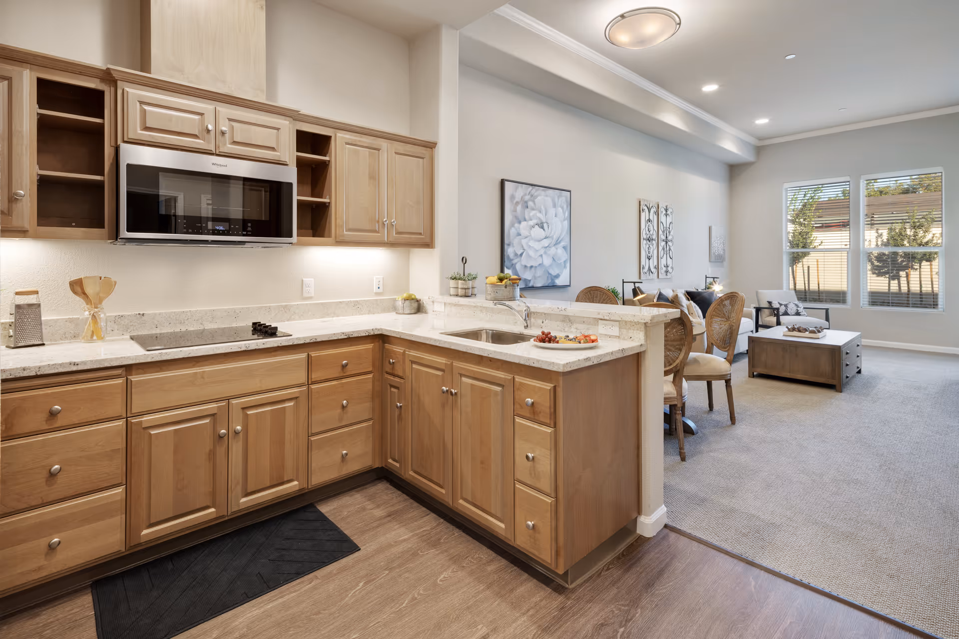 A modern kitchen with light wood cabinets, a built-in microwave, and a white countertop. The kitchen opens into a living area with carpeted flooring, a coffee table, chairs, and large windows letting in natural light. The walls are decorated with artwork and the space is well-lit with ceiling lights.