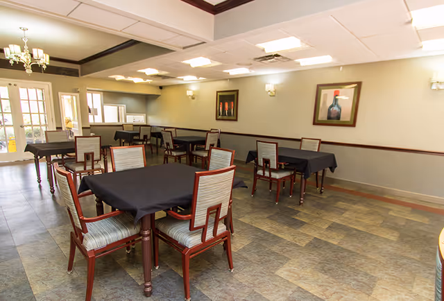 A dining room with several tables covered in black tablecloths and wooden chairs with striped cushions. The room has tiled flooring, light green walls with dark wood trim, framed artwork, and ceiling lights. There are glass doors leading outside on the left side.