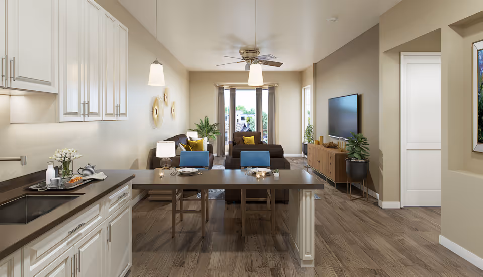A modern open-concept living space featuring a kitchen area with white cabinets and a dark countertop on the left, a dining table with two blue chairs in the center, and a living room with brown sofas, a ceiling fan, a large flat-screen TV on a wooden console, and large windows letting in natural light at the far end.