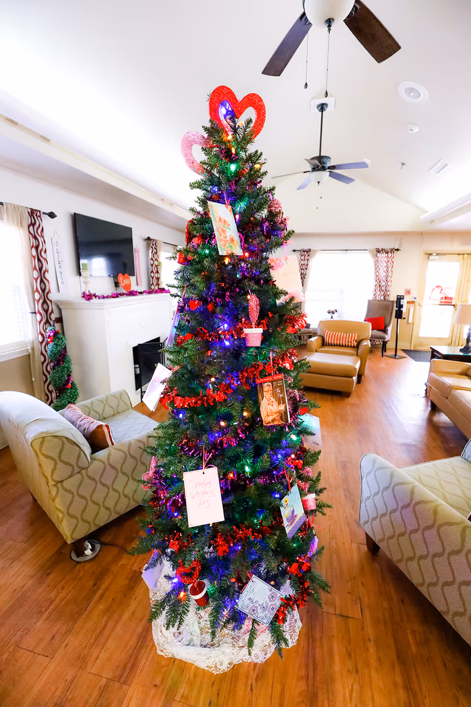 A decorated Christmas tree with colorful lights, red garlands, and various cards and ornaments stands in the center of a cozy living room. The room features wooden floors, patterned armchairs, a fireplace with a mounted TV above it, and large windows with patterned curtains allowing natural light to enter.