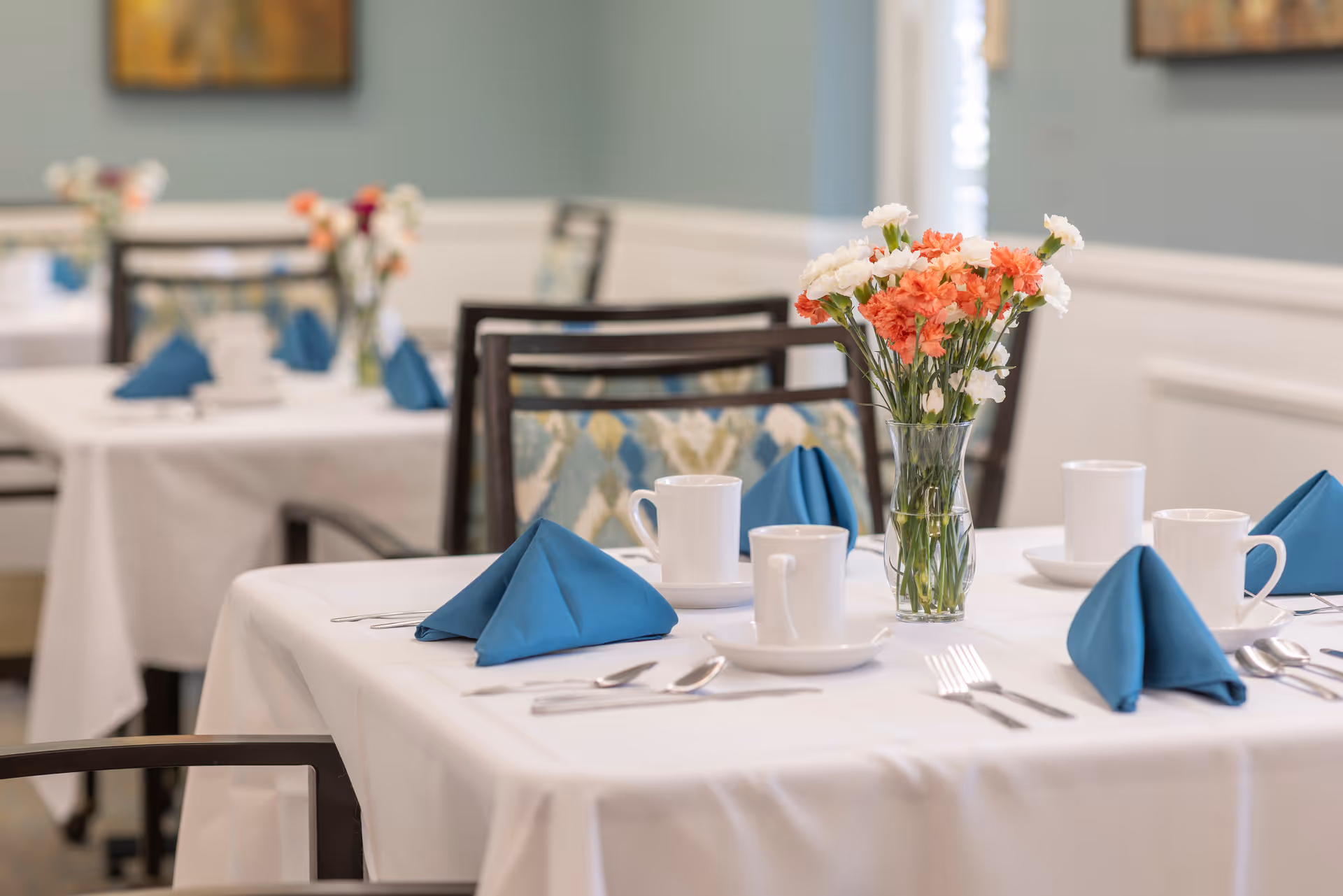 A dining table set with white tablecloth, blue folded napkins, white coffee cups on saucers, silverware, and a glass vase with orange and white flowers in a softly lit dining room.