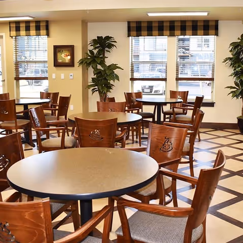 A bright dining area with several round tables and wooden chairs featuring a coffee cup design on the backrests. The room has large windows with checkered valances, potted plants, and a patterned floor.