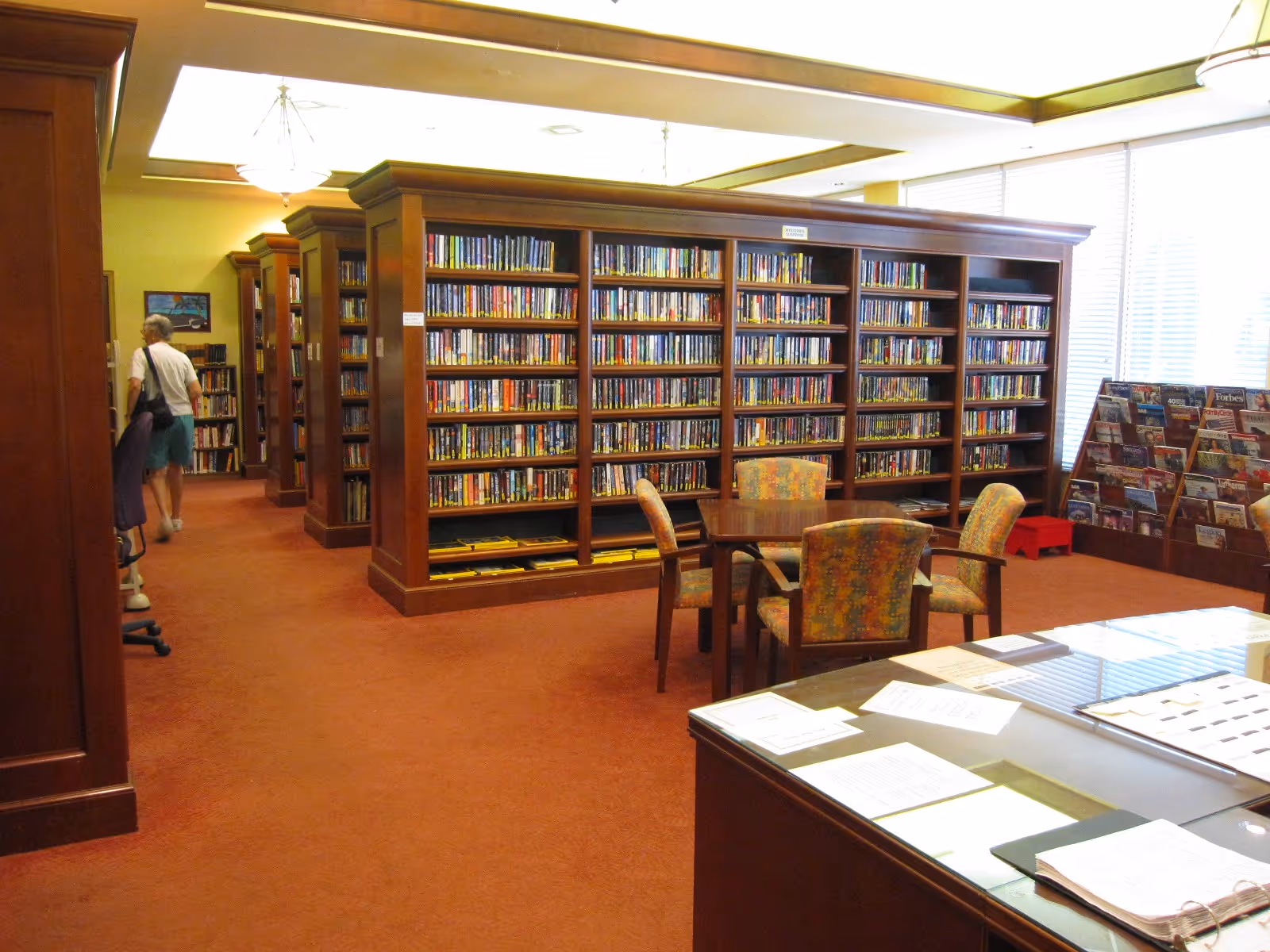 Interior of a library room with wooden bookshelves filled with books, a table with four patterned chairs in the center, a magazine rack near large windows with blinds, and a person walking away in the background.