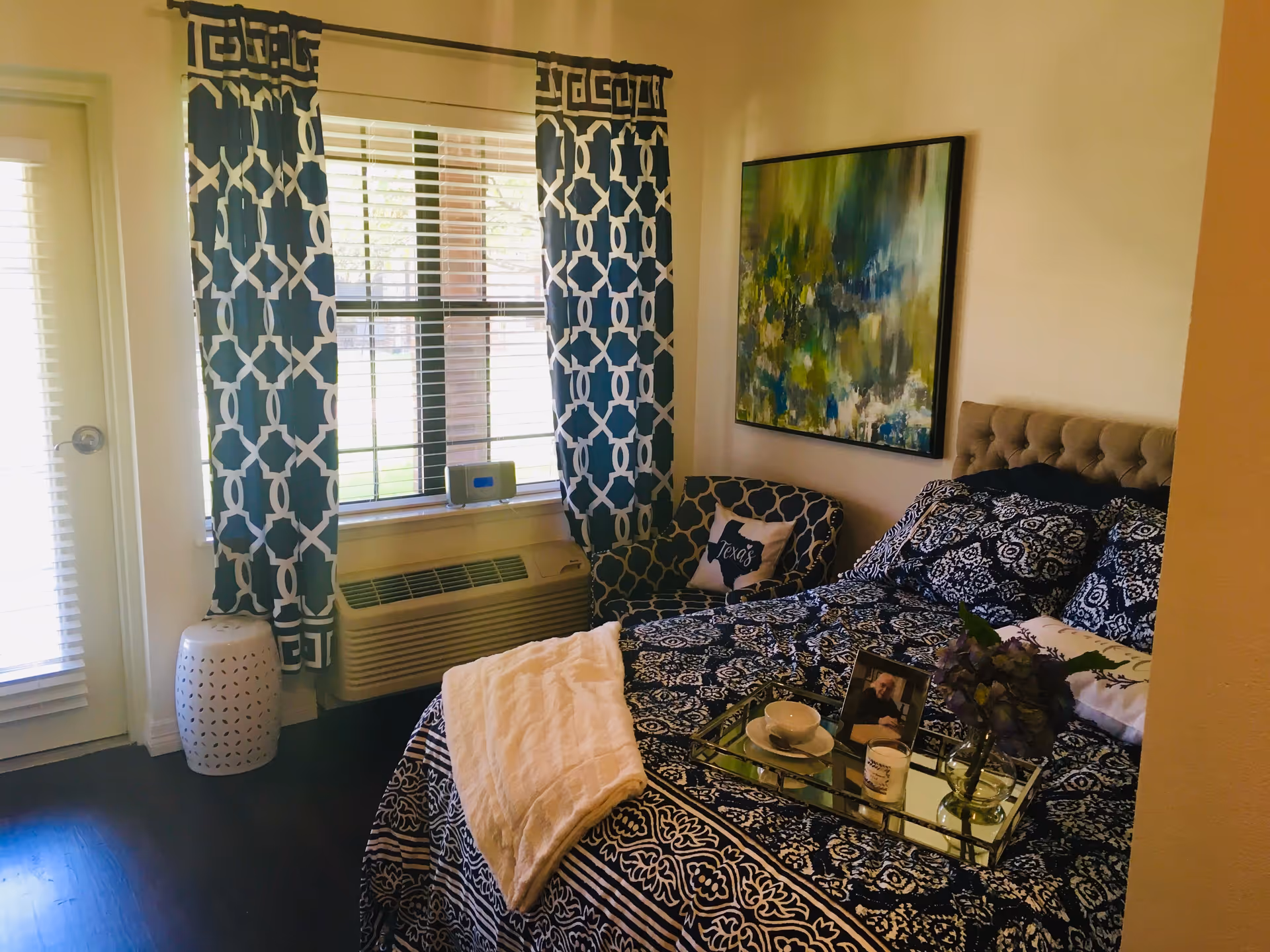 A cozy bedroom with a bed covered in a dark blue and white patterned comforter and pillows. A white throw blanket is draped over the bed. A tray on the bed holds a cup and saucer, a candle, a small vase with flowers, and a framed photo. Next to the bed is a patterned armchair with a pillow that says 'Texas'. The room has a window with white blinds and dark blue patterned curtains. There is a white ceramic stool near the window and an air conditioning unit below the window. A colorful abstract painting hangs on the wall above the armchair.