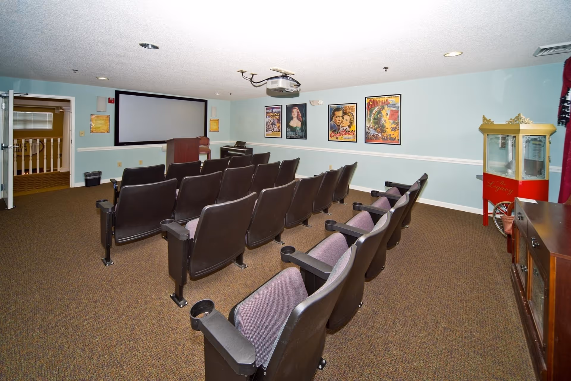A small movie theater room with two rows of black and purple cushioned seats facing a large projection screen. The walls are light blue with classic movie posters including Casablanca and Godzilla. A popcorn machine labeled 'The Legacy' is positioned on the right side of the room. There is a projector mounted on the ceiling and an open door leading to a hallway.
