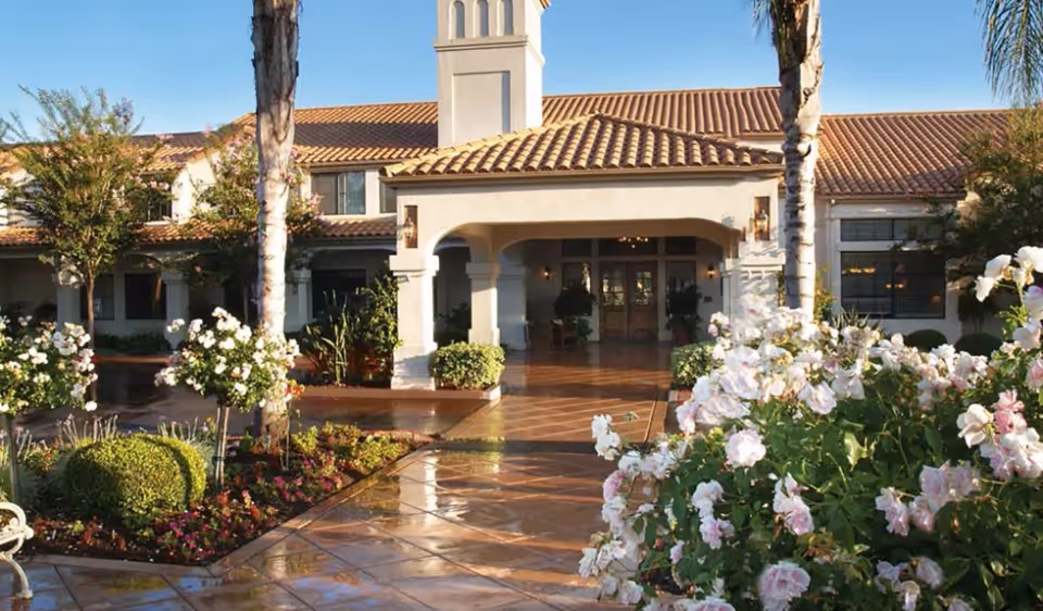 Front entrance of Oakmont of Escondido Hills facility with a tiled walkway, palm trees, flowering bushes, and a building with a tiled roof and arched entryway.