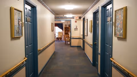 Carpeted interior hallway in a memory care facility with blue doors, handrails, framed artwork, and a small sitting area visible at the end.