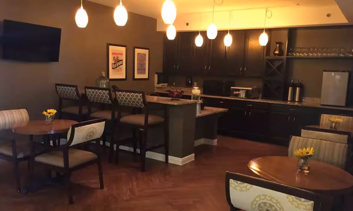 Interior view of a senior living facility's dining and kitchen area with round wooden tables and chairs, a kitchen counter with bar stools, pendant lights hanging from the ceiling, dark cabinetry, and small flower vases on the tables.