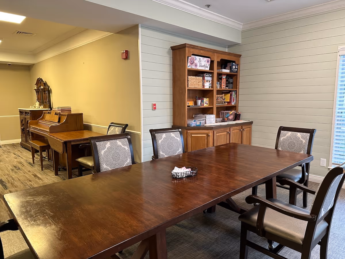 Communal dining/activity room with a long wooden table and chairs, a cabinet with books and games, and a piano along the wall.