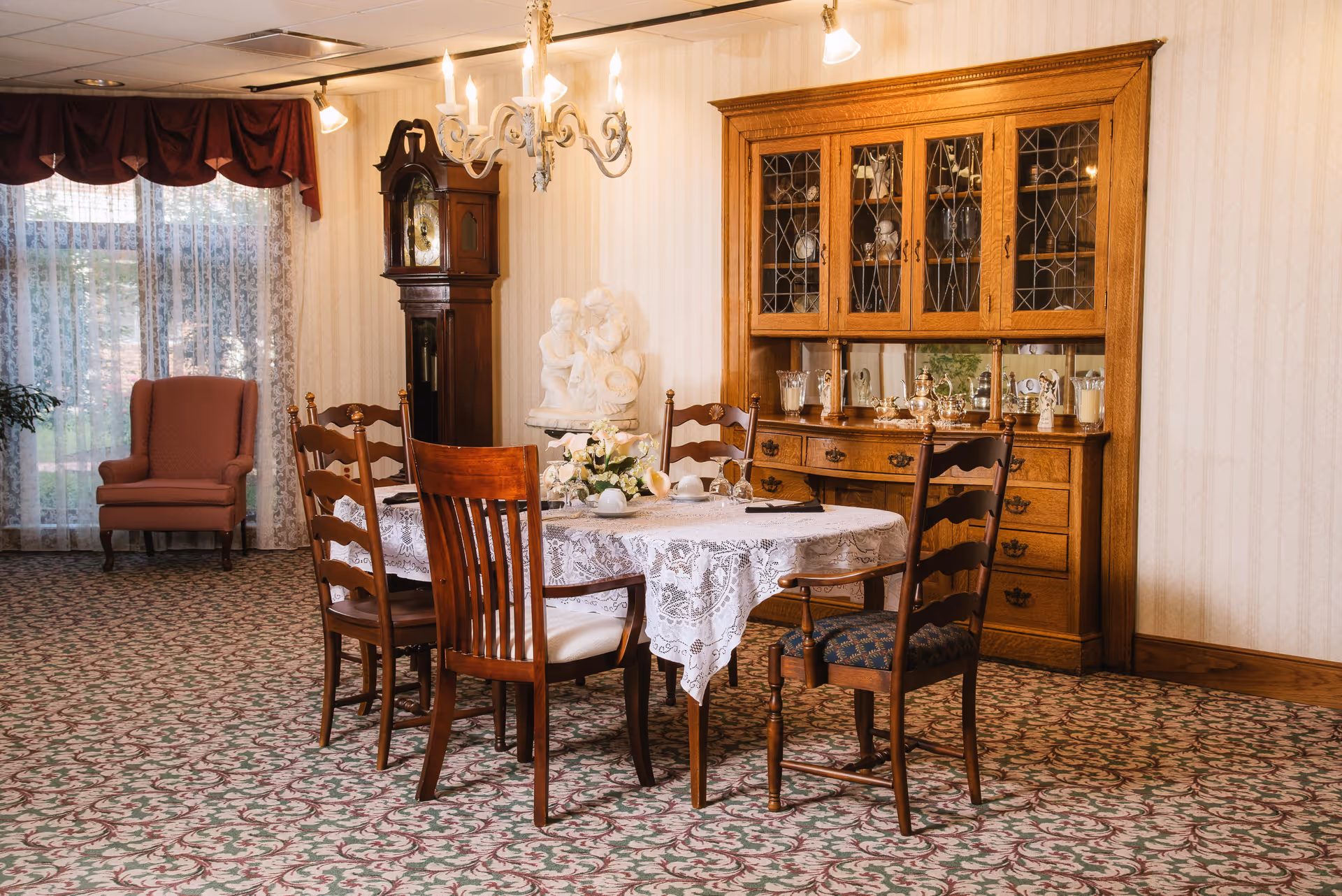 A traditionally furnished dining room with a lace-covered table and wooden chairs, a china cabinet, grandfather clock, and an upholstered armchair by a window.