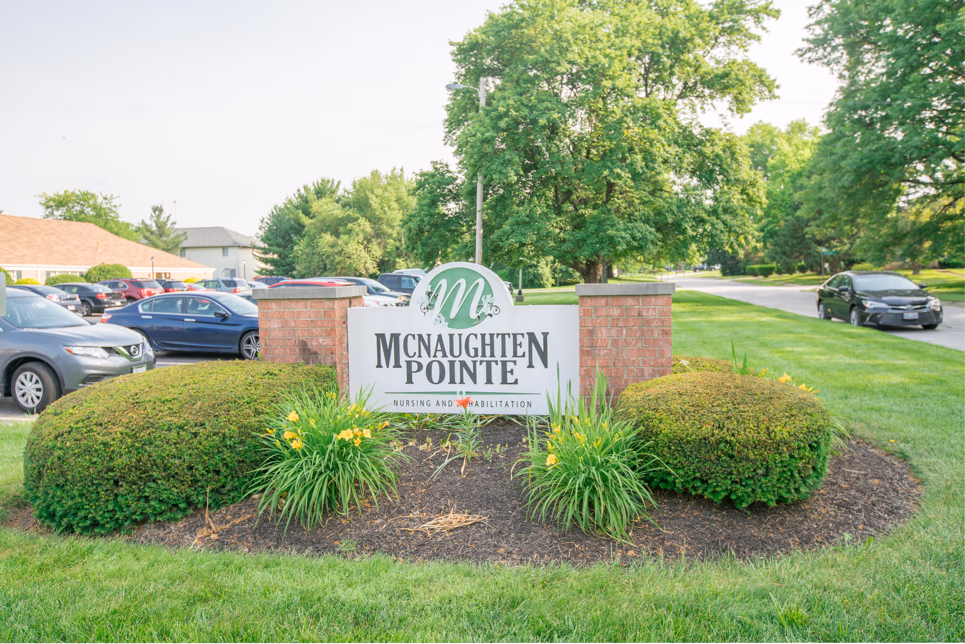 Entrance sign reading "McNaughten Pointe" set in landscaped greenery with parked cars and trees in the background.