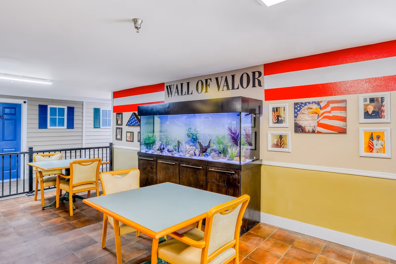 Interior of a senior living facility featuring a seating area with tables and chairs. A large fish tank is mounted on a dark wooden cabinet against a wall painted with red and white stripes and the words 'WALL OF VALOR'. The wall also displays framed pictures, including an American flag and a bald eagle painting.