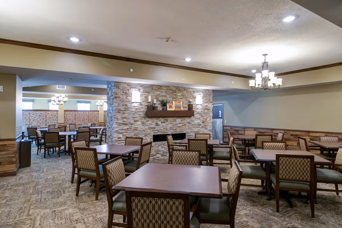A spacious dining room in a senior living facility featuring multiple square wooden tables with patterned cushioned chairs. The room has a stone accent wall with a mantle decorated with candles and picture frames. Ceiling lights and a chandelier provide illumination, and the carpet has a textured pattern. The walls are painted in neutral tones with wood paneling along the lower half.
