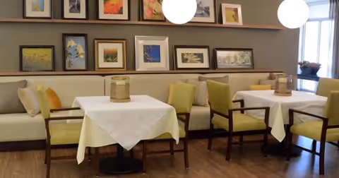 A dining area with tables covered in white tablecloths, surrounded by green cushioned chairs and a long beige bench along the wall. The wall is decorated with multiple framed pictures and two round hanging light fixtures. A window with blinds and a small plant on the windowsill is visible on the right side.