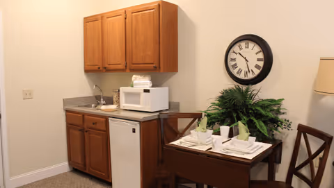 Small kitchenette area with wooden cabinets, a countertop with a sink, a microwave, and a mini refrigerator. Next to the kitchenette is a small dining table set for two with plates, glasses, napkins, and a green plant centerpiece. A round wall clock with Roman numerals hangs above the table, and a floor lamp is partially visible on the right side.