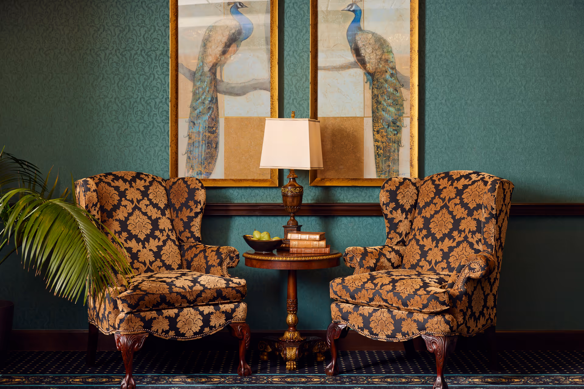Two ornate wingback chairs flanking a round side table with a lamp and peacock artwork on a green patterned wall.