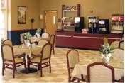 Dining area with several tables covered with beige tablecloths and floral centerpieces. Chairs with cushioned seats surround the tables. In the background, there is a beverage station with coffee and soda machines against a wall, and a door to the left side.