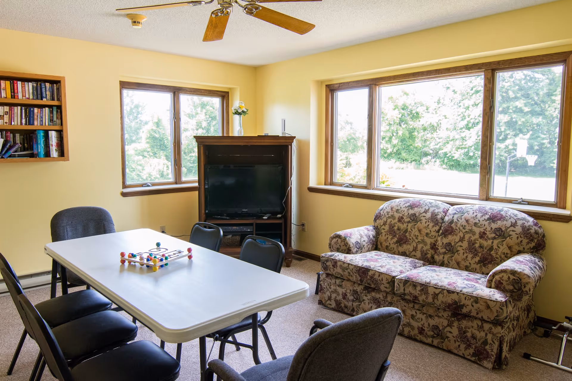 A bright room with yellow walls featuring a white rectangular table surrounded by black chairs, a floral patterned couch, a wooden cabinet with a TV, and large windows showing green trees outside. A bookshelf with books is mounted on the wall.