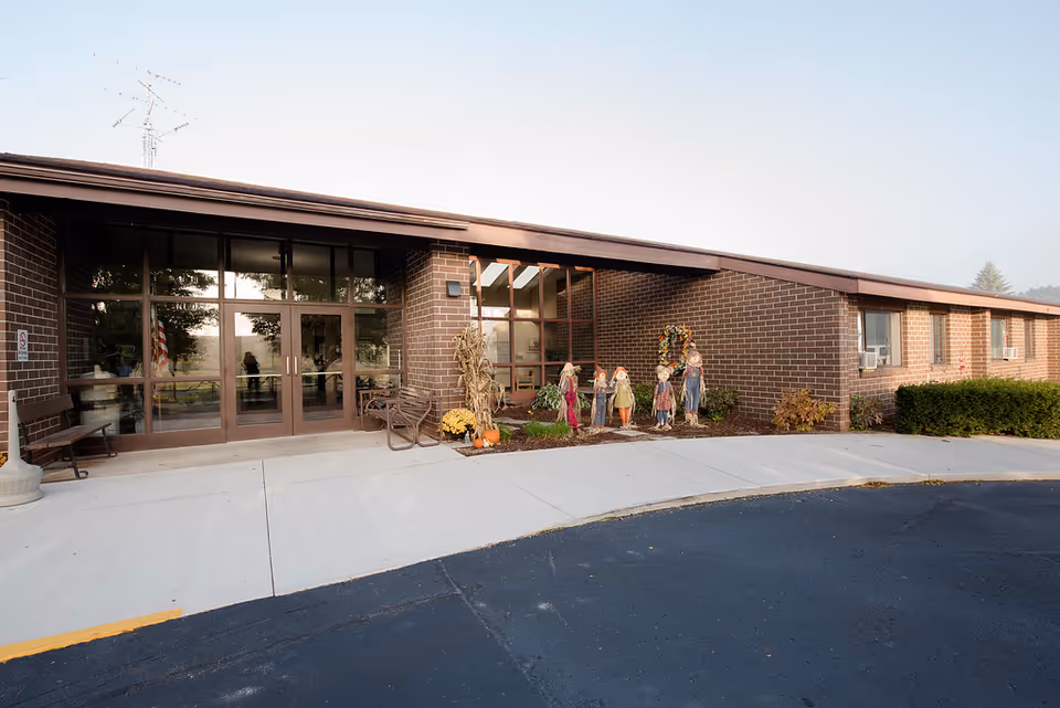 Exterior view of a single-story brick building with large glass doors and windows. There are fall decorations including scarecrows, pumpkins, and a wreath near the entrance. A bench is placed near the door, and the pavement and parking area are visible in front.