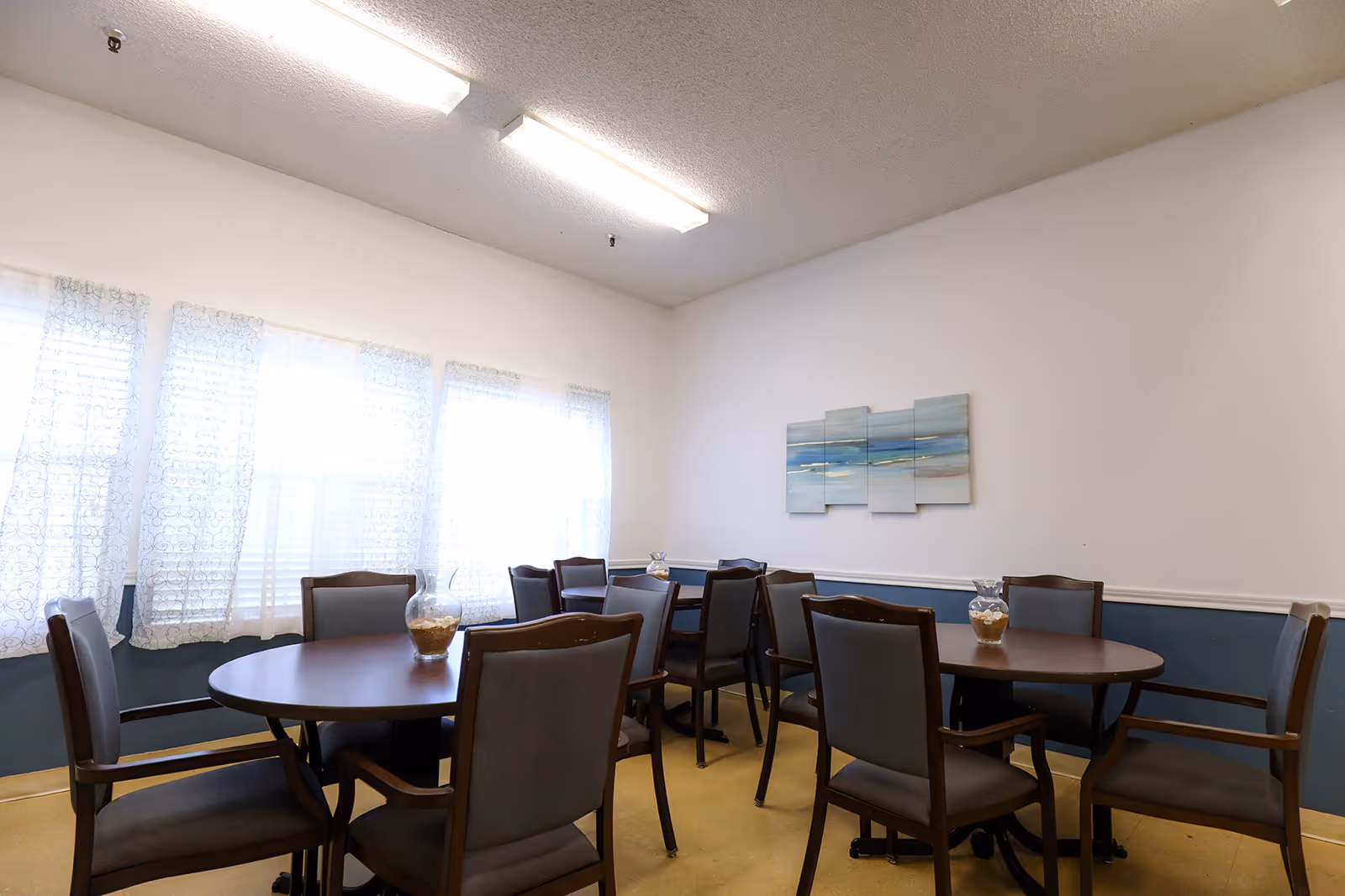 A dining room with two round wooden tables, each surrounded by six cushioned chairs. The room has large windows with sheer white curtains allowing natural light to enter. On the wall, there is a multi-panel abstract painting with blue and beige tones. The walls are painted white with a blue wainscoting, and the ceiling has fluorescent lighting.